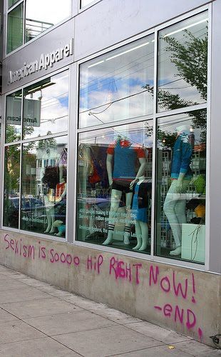 an American Apparel storefront with spraypainted pink graffiti on the front that says Sexism is soooo hip right now! -end.
