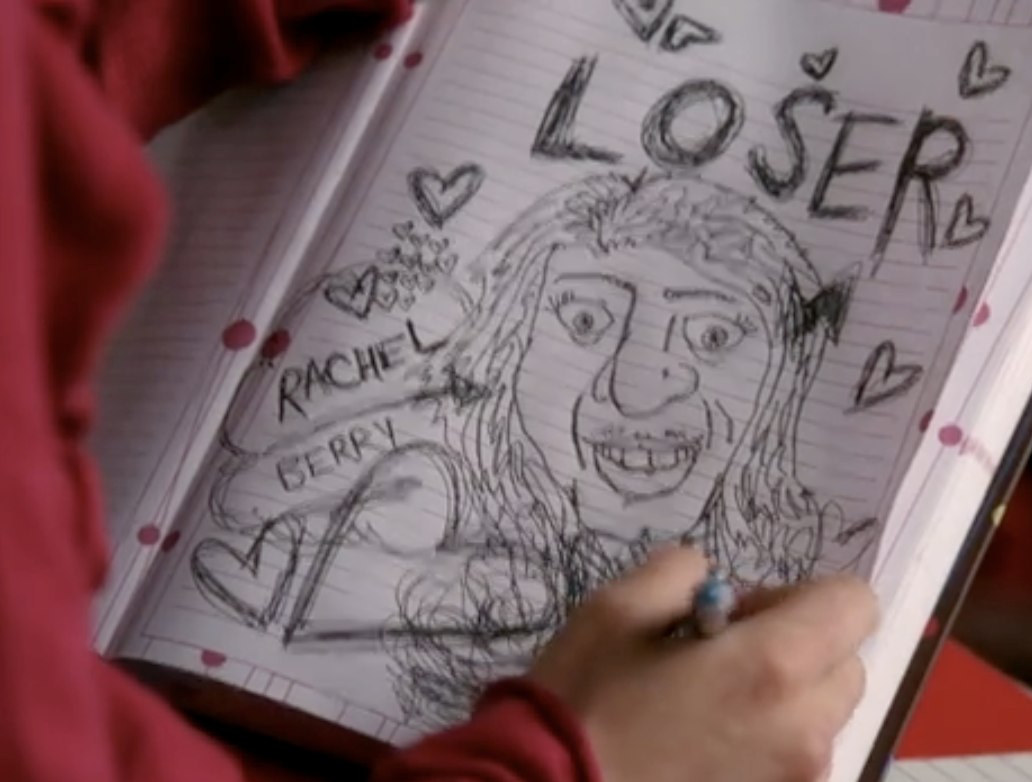 a woman's hand scrawls the word 'Loser' on a high school notebook with a stereotypical drawing of a jewish woman, with frizzy hair, a large nose, etc and the character's name, Rachel Berry, written underneath it.