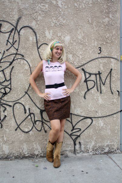 Courtney wearing an amazing blonde wig, leaning against a wall smiling