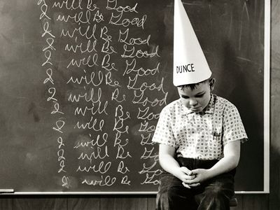Sad looking child in a dunce-cap sitting in front of a blackboard