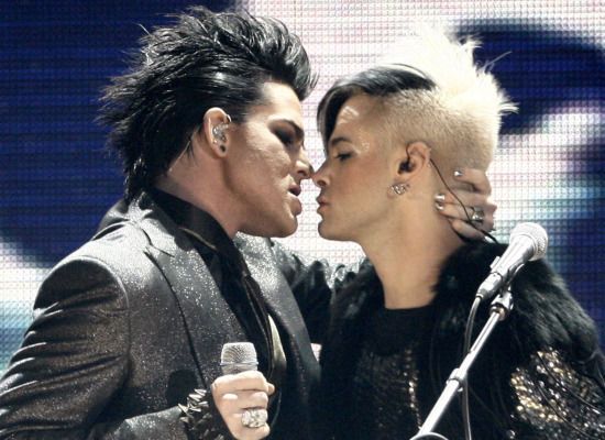 Adam Lambert, all glammed up, holds the back of a male band member's neck, going in for a kiss